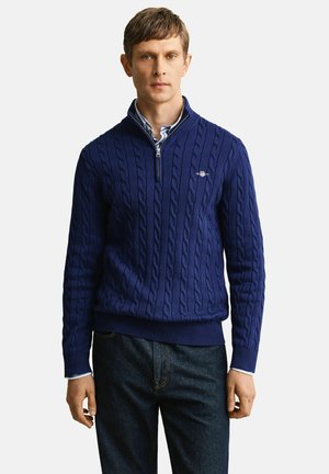 Mann trägt einen blauen, grob gestrickten Pullover mit Viertel-Reißverschluss über einem hellblauen Hemd mit Kragen und dunklen Jeans, steht vor einem einfarbigen Hintergrund.