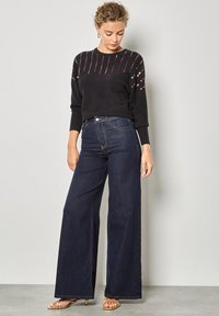 Pull noir avec des broderies colorées sur les épaules, associé à un jean évasé bleu foncé taille haute. Mannequin portant des sandales dorées à lanières.