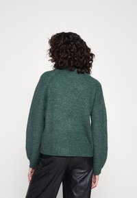 Femme aux cheveux bouclés portant un pull en tricot vert foncé et un pantalon en cuir noir, vue de dos sur un fond blanc uni.