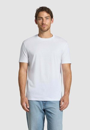 Uomo con capelli castani corti che indossa una maglietta bianca semplice a maniche corte e jeans azzurri, in piedi davanti a uno sfondo semplice.