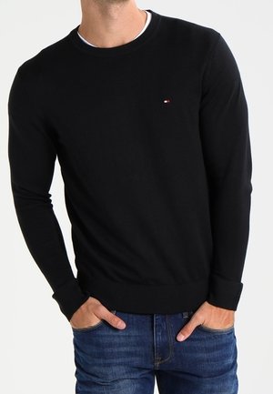 Man wearing a black long-sleeve crewneck sweater with a small red, white, and blue logo on the chest, paired with blue jeans.