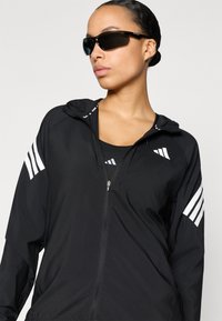 Chaqueta negra con cremallera y capucha, con franjas blancas como detalle en las mangas; combinada con gafas de sol oscuras. Tejido liso y ligero.