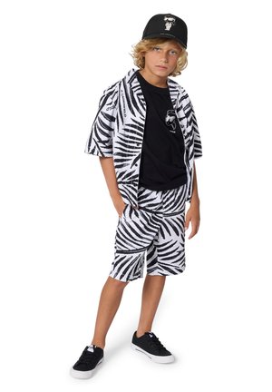 Blond chłopiec w czarnej czapce, czarnej koszulce, koszuli w paski zebra i pasujących szortach oraz czarnych tenisówkach, stojący z rękami w kieszeniach.