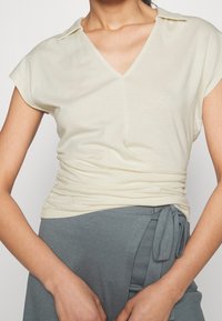 Light yellow short-sleeve top with a V-neck and collar, featuring a gathered side seam. Styled with a gray wrap skirt, showing a tied detail.