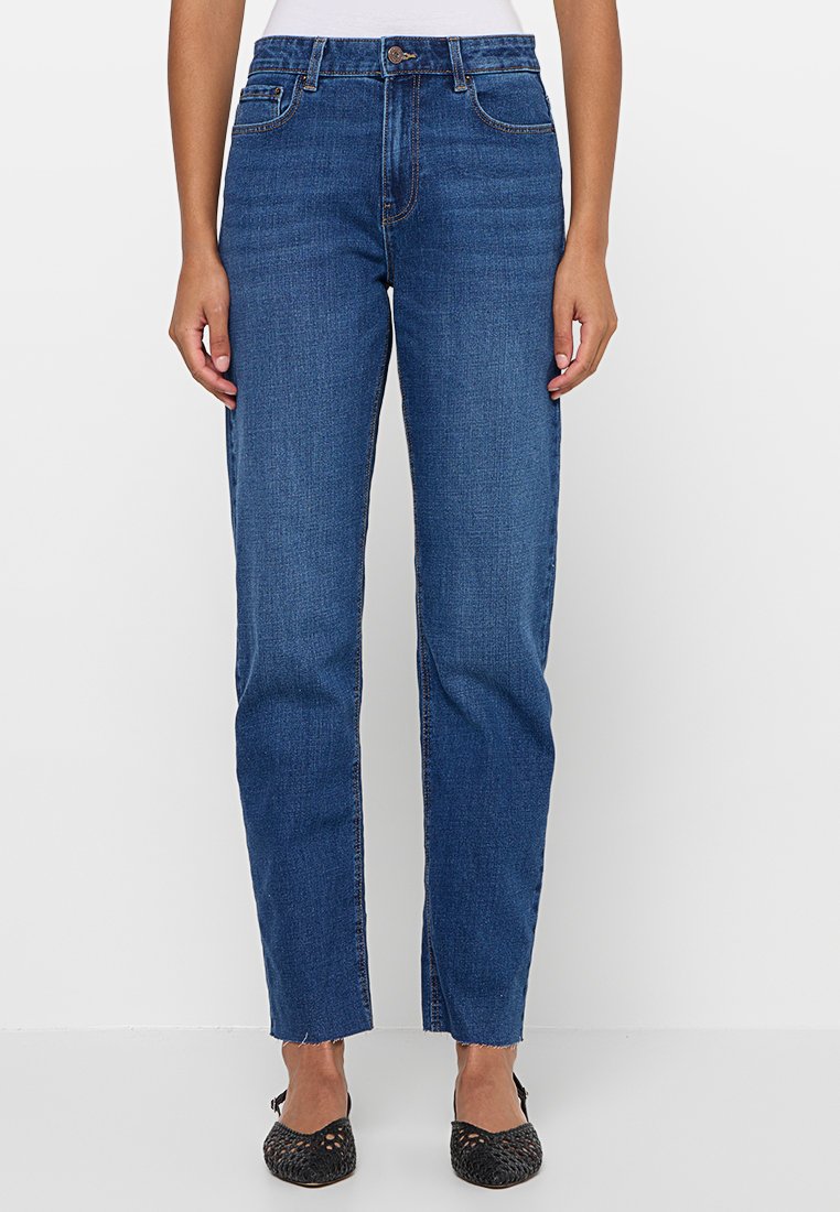 Blaue Jeans aus Denim mit geradem Bein, ausgestattet mit einem Knopfverschluss, fünf Taschen und umsäumten Säumen. Tragen mit schwarzen Ballerinas.