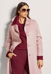 Helle rosa Jacke mit strukturiertem Finish, großem Kragen und zwei Knöpfen. Darüber eine burgunderfarbene Bluse und passende Hose. Dunkle Sonnenbrille als Akzent.
