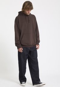 Sweatshirt à capuche marron avec une poche avant, texte de logo noir. Associé à un jean bleu foncé ample et des baskets noires.