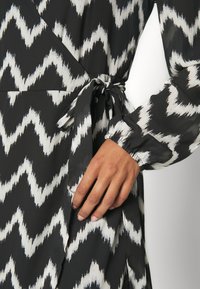 Main reposant sur la hanche d'une robe portefeuille noire et blanche à motif zigzag, avec une manche longue bouffante et une ceinture en tissu à la taille.