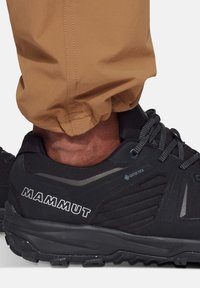 Zwarte Mammut-schoenen beschikken over een textuur bovenwerk met GORE-TEX-merktekens, een gevoerde zool en een strak ontwerp, gedragen met een paar bruine broeken.