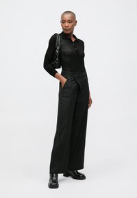 Camisola preta de manga longa com botões, combinada com calças largas pretas com riscas e sapatos pretos robustos. Os acessórios incluem uma bolsa preta.