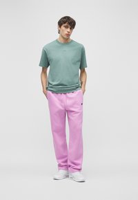 Mladý muž oblečený v svetlozelenej tričku Nike, svetloružových teplákoch Nike, bielych teniskách, stojaci s rukami v kapse na jednoduchom pozadí.