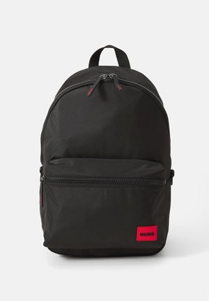 Black fabric backpack with front zip pocket, dual zipper closure, top handle, and red rectangular label reading "HUGO" in bottom right corner.