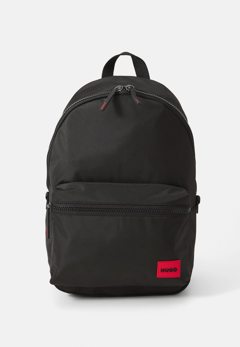 Mochila de tela negra con bolsillo frontal con cremallera, cierre de doble cremallera, asa superior y etiqueta rectangular roja con la inscripción "HUGO" en la esquina inferior derecha.