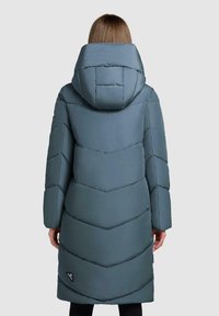 Kvindelig i en lang, polstret blå-grå vinterfrakke med en stor hætte, stående med ryggen til kameraet mod en ensfarvet baggrund.