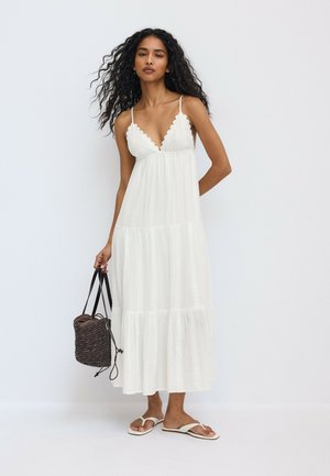 Femme aux longs cheveux bouclés portant une robe midi blanche sans manches et des sandales blanches, tenant un sac à main tressé foncé.