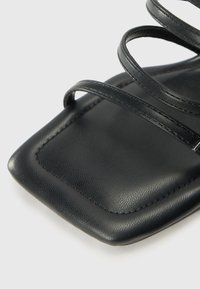 Sandale en cuir noir avec un design à bout carré, ornée de plusieurs fines sangles entrecroisées. Texture lisse et détails de surpiqûres subtils.