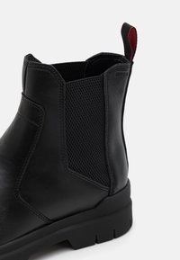 Bottine en cuir noir avec panneau latéral élastique, détails de couture et une languette en tissu rouge et noir à l'arrière sur fond blanc.