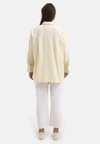 Camicia sopra misura gialla a righe con linee multicolori verticali, maniche lunghe e orlo curvo, abbinata a pantaloni bianchi a gamba larga.