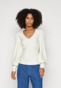 Long-sleeve cream top with a V-neck and gathered detailing at the center. Features puffed sleeves and a fitted silhouette.
