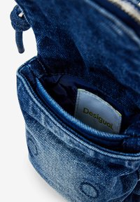 Denimtasche mit strukturierter Oberfläche, ausgestattet mit einem Reißverschluss oben, in dunkler blauer Farbe, mit innerem Futter und einem sichtbaren weißen Etikett mit "Desigual."