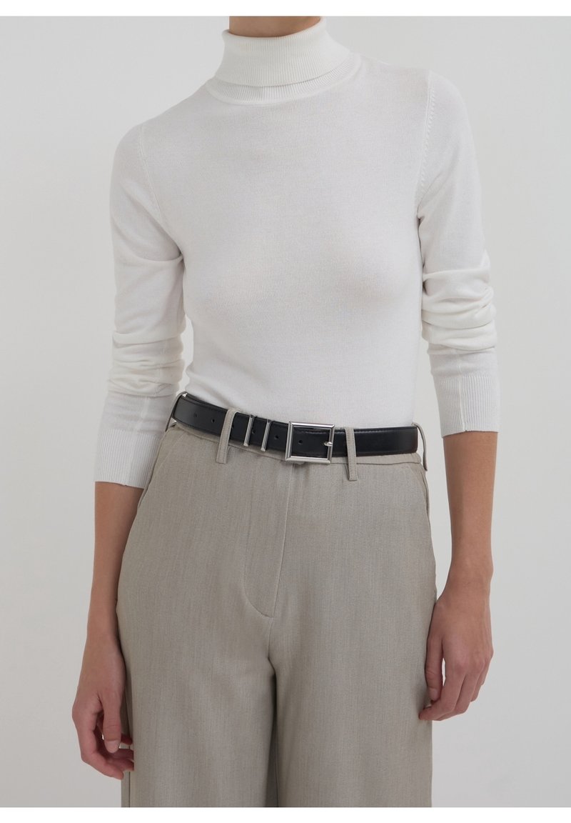 Femme portant un pull à col roulé blanc à manches longues, rentré dans un pantalon beige avec une ceinture noire et une boucle argentée.