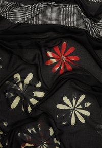 Écharpe noire à motifs avec des dessins floraux en rouge et crème. Présente une texture douce et fluide avec un accent pied-de-poule en haut.