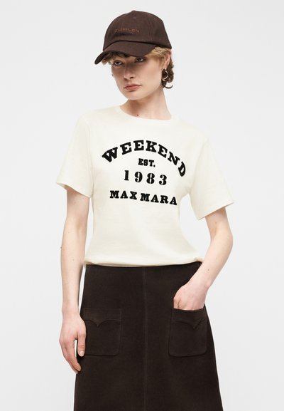 Νεαρή γυναίκα με λευκό μπλουζάκι με την επιγραφή "WEEKEND EST. 1983 MAX MARA", σκούρα φούστα με μπροστινές τσέπες, καφέ καπέλο και χρυσούς κρίκους στα αυτιά.