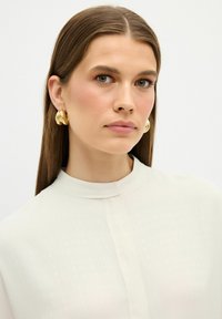 Gouden creolen, glad en gepolijst, gecombineerd met een hoge-neklijng, crèmekleurige blouse met een subtiele textuur en een centrale knopenlijst.