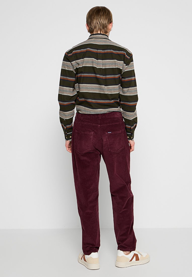 Maroonowe spodnie kordowe z tylnymi kieszeniami, połączone z koszulą w paski w odcieniach zieleni i brązu zapinaną na guziki. Całość uzupełniają jasne sneakersy.