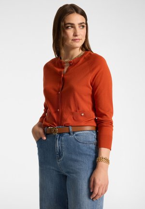 Femme portant un cardigan orange boutonné avec des poches devant, un jean bleu, une ceinture marron avec une boucle dorée et des bijoux dorés regardant vers la droite.