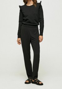 Sudadera negra con hombros fruncidos, combinada con pantalones negros y sandalias de tiras. Tejido suave con un corte relajado y diseño minimalista.