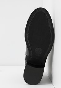 Chaussure en cuir noir avec un dessus lisse, un bout arrondi et une semelle texturée. Présente des détails cousus et une marque sur la semelle.