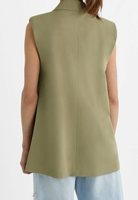 Blazer sans manches vert olive avec col pointu et dos structuré. Tissu lisse avec une coupe ajustée et une longueur moyenne.