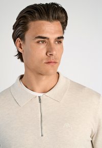 Beige zippered polo shirt med ribbad textur, med en mjuk krage och liten logotyp på bröstet. Modellen har kort hår och ett neutralt uttryck.