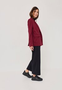 Burgundijas žakete ar divām priekšējām kabatām, apvienota ar melniem plašajiem biksēm un melniem sporta apaviem. Gluda auduma, strukturēts dizains.