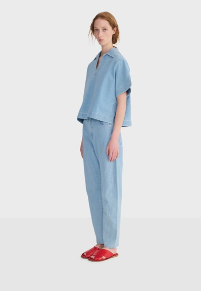 Mujer de pie con blusa de mezclilla azul claro de manga corta y pantalones, combinados con sandalias planas rojas de punta abierta, sobre un fondo blanco liso.