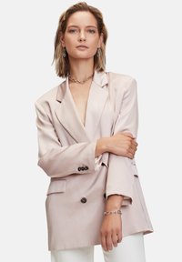 Blazer in satin rosa chiaro con scollo a V profondo, bottoni doppi e tasche laterali; abbinato a un bracciale a catena argentato e pantaloni bianchi.