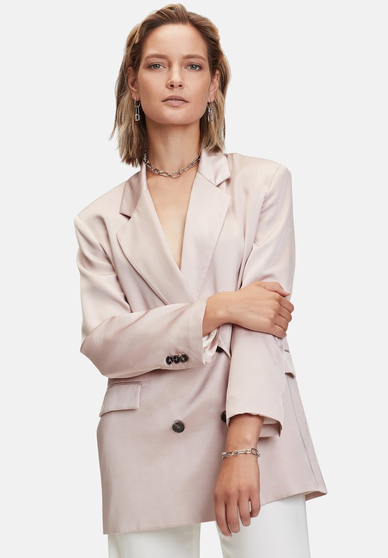 Blazer in satin rosa chiaro con scollo a V profondo, bottoni doppi e tasche laterali; abbinato a un bracciale a catena argentato e pantaloni bianchi.