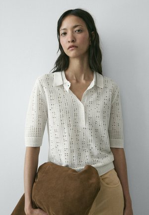 Junge Frau mit langen dunklen Haaren trägt ein weißes kurzes Strickoberteil und beige Hosen und hält eine große braune Wildleder-Clutch vor hellem Hintergrund.