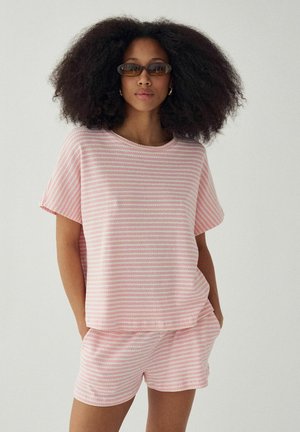 Kvinde med fyldigt krøllet hår iført solbriller og en pink- og hvidstribet kortærmet top samt matchende shorts, hænderne i lommerne.