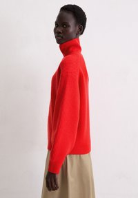 Roter gerippter Rollkragenpullover mit langen Ärmeln, locker geschnitten und mit strukturierter Strickoptik. Kombiniert mit einem beigen Rock vor hellem Hintergrund.