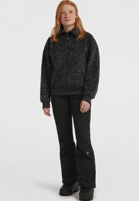 Vrouw met lang rood haar draagt een zwarte gebreide fleece trui, zwarte broek en zwarte laarzen, staand tegen een effen lichte achtergrond.