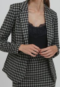 Kobieta w szaro-czarnej w kratę marynarce i spódnicy, z czarną bluzką z koronkowym wykończeniem, z rękami złożonymi przy talii, na neutralnym tle.