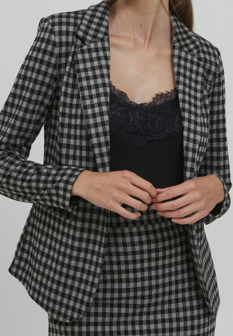 Kobieta w szaro-czarnej w kratę marynarce i spódnicy, z czarną bluzką z koronkowym wykończeniem, z rękami złożonymi przy talii, na neutralnym tle.