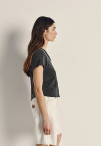 Ženska z dolgimi rjavimi lasmi, oblečena v črno kratko srajco in bele kratke hlače do kolen, stoji pokonci ob preprosti steni.