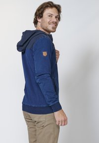 Navy blue hoodie with ribbed hood and cuffs, featuring a leather shoulder patch, kangaroo pocket, and contrasting textured shoulder panels.