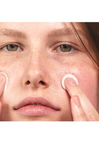 Närbild av ett ansikte som applicerar en vit kräm med fingrarna. Huden ser återfuktad ut med en slät textur och en lätt lyster. Fräknar syns på kinderna.