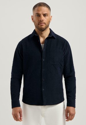 Dunkelblauses Langarmhemd mit einem strukturierten Muster, einem button-down Kragen und einer einzelnen Brusttasche; kombiniert mit hellen Hosen.