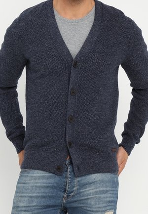 Dunkelblauer Cardigan mit V-Ausschnitt, strukturiertem Strick, fünf schwarzen Knöpfen und gerippten Bündchen. Über einem grauen Hemd getragen und mit ausgewaschenen Jeans kombiniert.