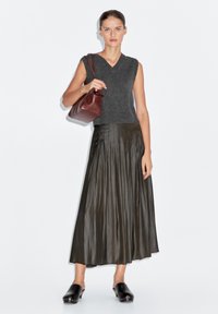 Top grigio senza maniche in maglia abbinato a una gonna verde scuro plissettata e zoccoli neri. La modella tiene una borsa marrone. Sfondo minimalista.